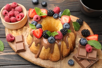 Photo of Tasty Bundt cake with berries, chocolate, mint and coffee on wooden table, closeup