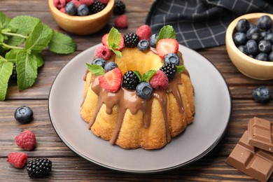 Photo of Tasty Bundt cake with berries, chocolate and mint on wooden table, closeup