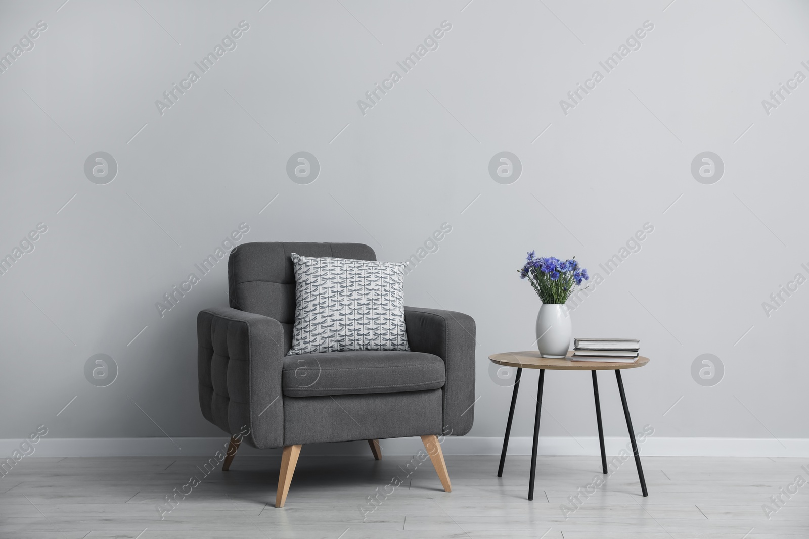 Comfortable armchair, vase with flowers and books near light gray wall indoors Photo of Comfortable armchair, vase with flowers and books near light gray wall indoors