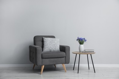 Photo of Comfortable armchair, vase with flowers and books near light gray wall indoors