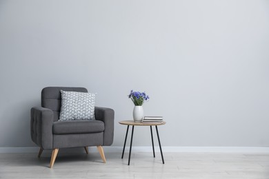 Photo of Comfortable armchair, vase with flowers and books near light gray wall indoors. Space for text