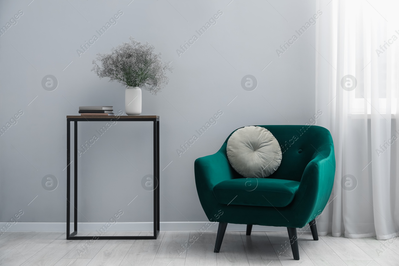 Photo of Comfortable armchair, vase with flowers and books near light gray wall indoors