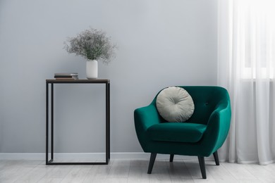 Photo of Comfortable armchair, vase with flowers and books near light gray wall indoors