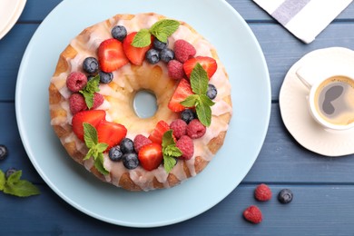 Photo of Delicious bundt cake with berries, glaze, mint and cup of coffee on blue wooden table, flat lay