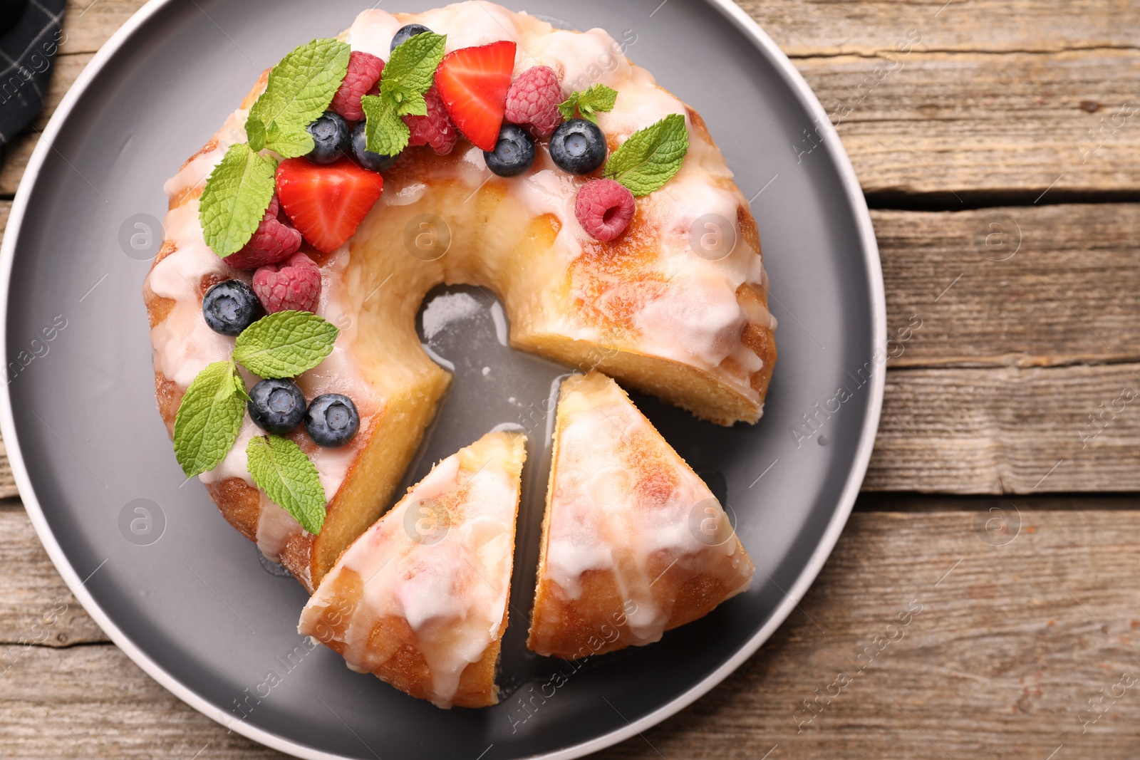 Photo of Pieces of delicious bundt cake with berries, glaze and mint on wooden table, top view