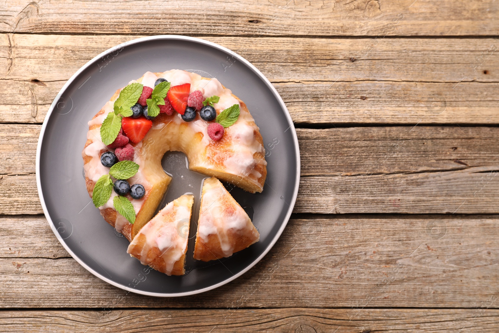 Photo of Pieces of delicious bundt cake with berries, glaze and mint on wooden table, top view. Space for text