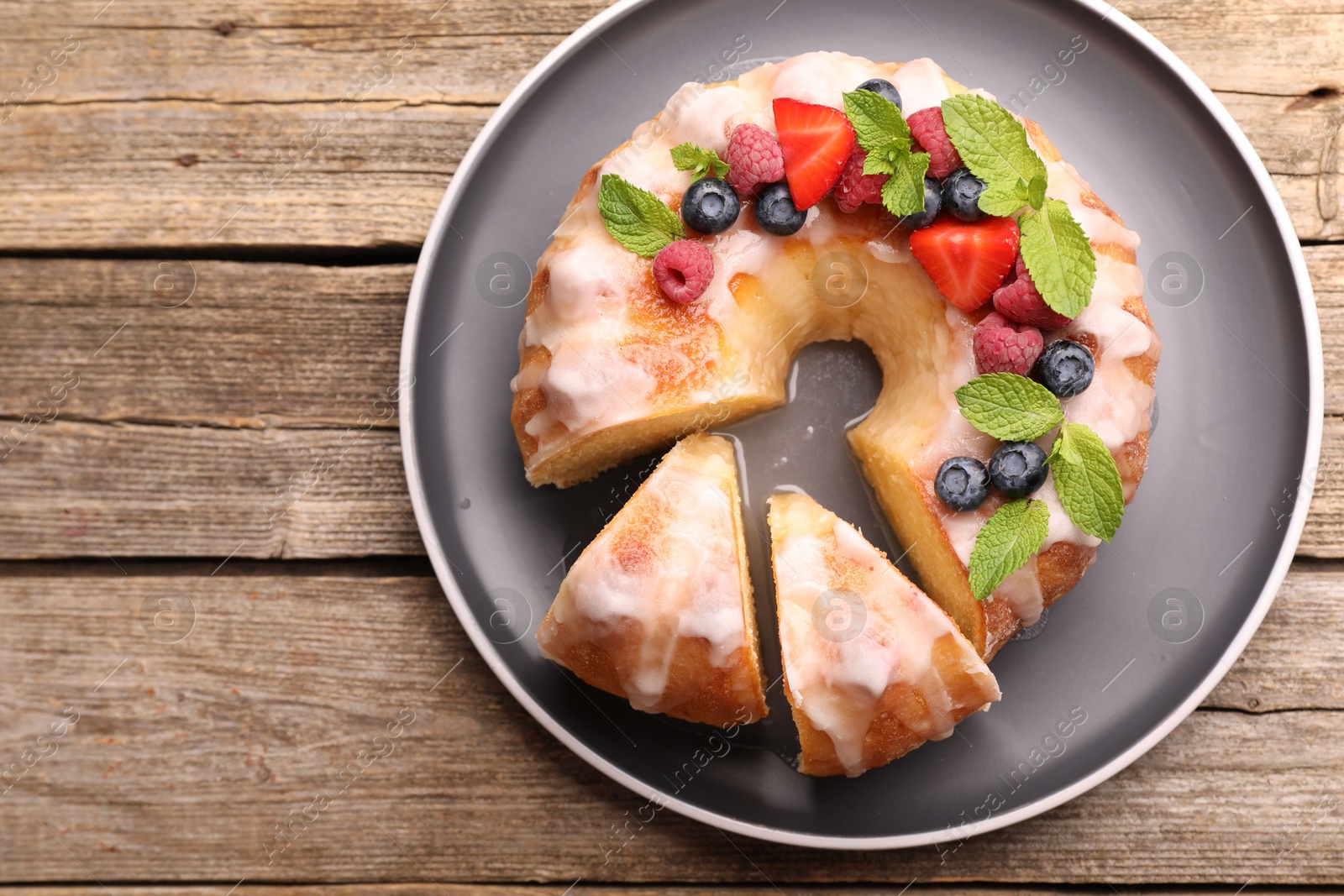 Photo of Pieces of delicious bundt cake with berries, glaze and mint on wooden table, top view