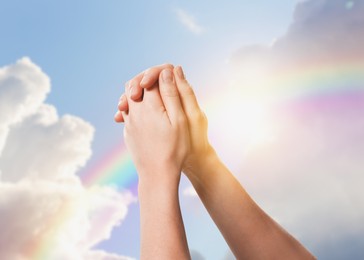 Image of Hope. Woman praying holding hands against blue sky with sun and rainbow, closeup