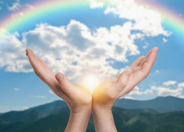 Image of Hope. Man holding hands against sky with rainbow and sun in mountains, closeup