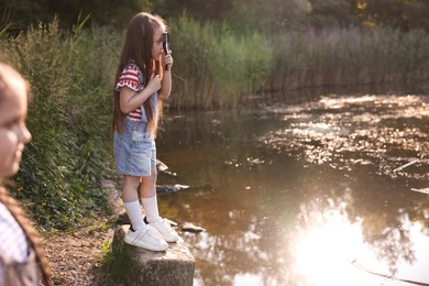 Photo of Cute little girl looking through magnifying glass by lake in nature