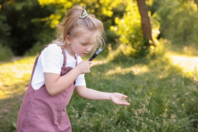 Photo of Cute little girl looking at plant through magnifying glass in nature