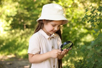Photo of Cute little girl looking at branch with green leaves through magnifying glass in nature