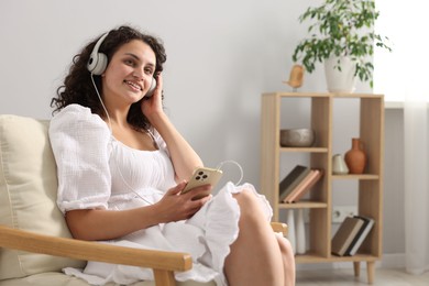 Photo of Young woman with headphones and smartphone listening to music on armchair at home
