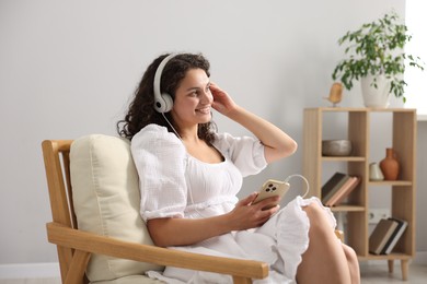 Photo of Young woman with headphones and smartphone listening to music on armchair at home