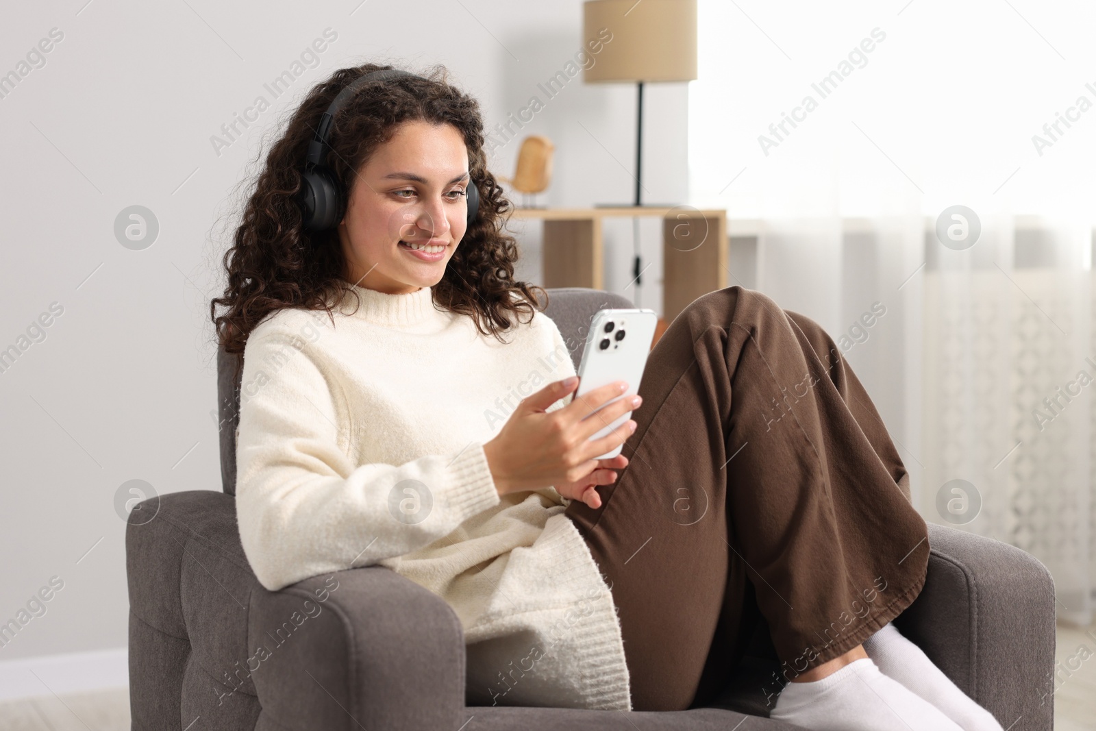 Photo of Young woman with headphones and smartphone listening to music on armchair at home