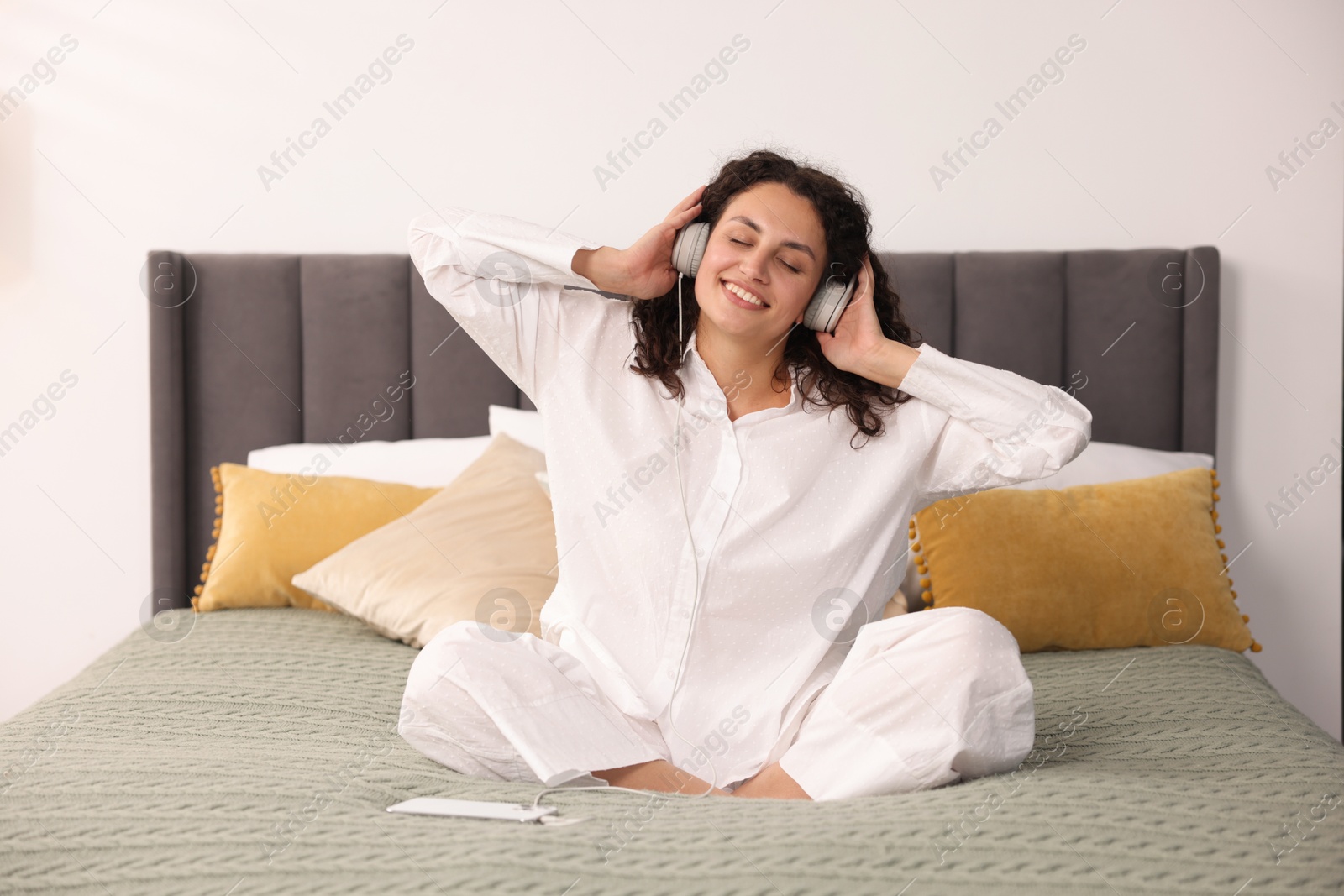Photo of Young woman with headphones and smartphone listening to music on bed at home