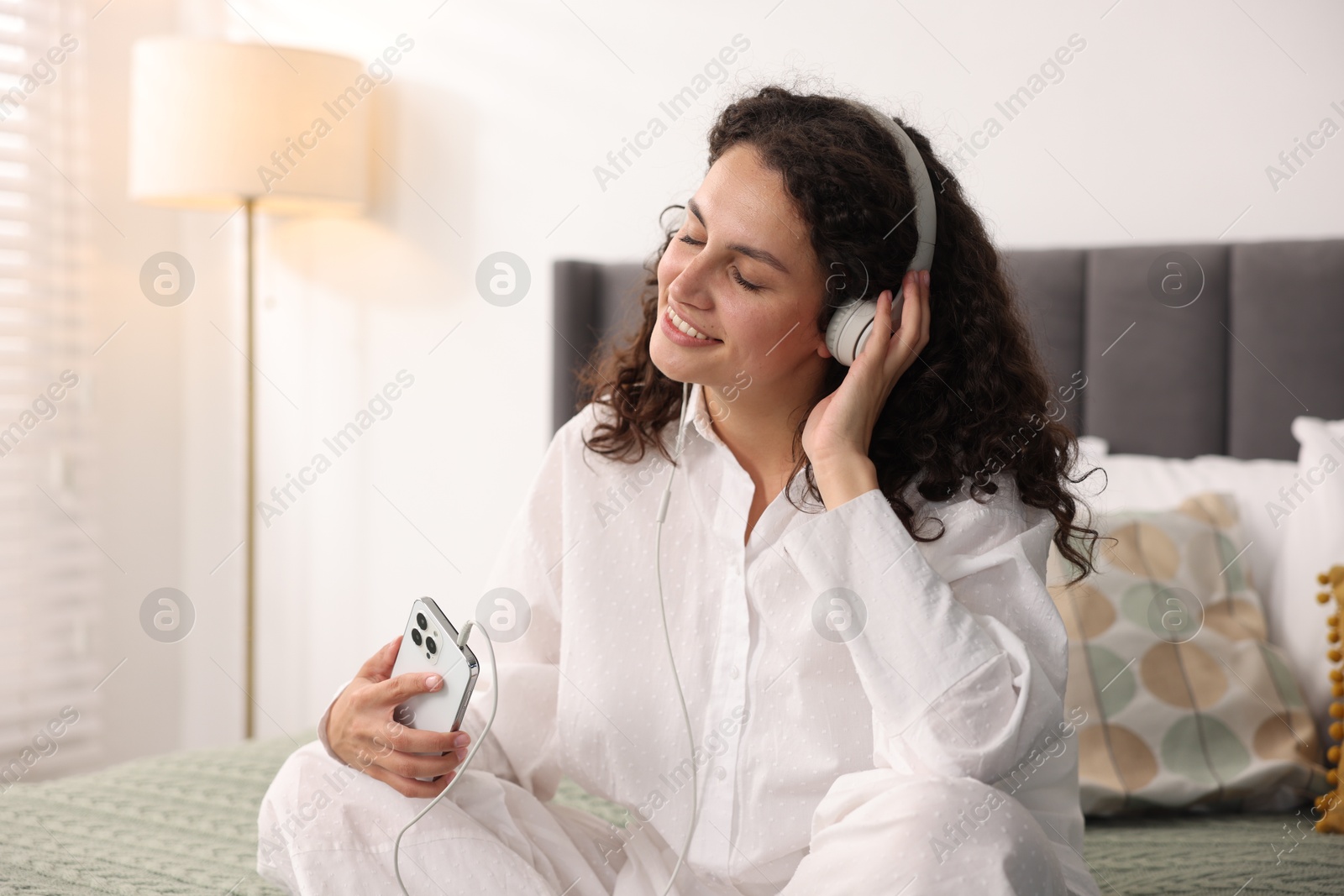 Photo of Young woman with headphones and smartphone listening to music on bed at home