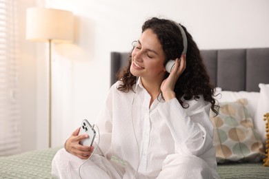 Photo of Young woman with headphones and smartphone listening to music on bed at home