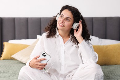 Photo of Young woman with headphones and smartphone listening to music on bed at home