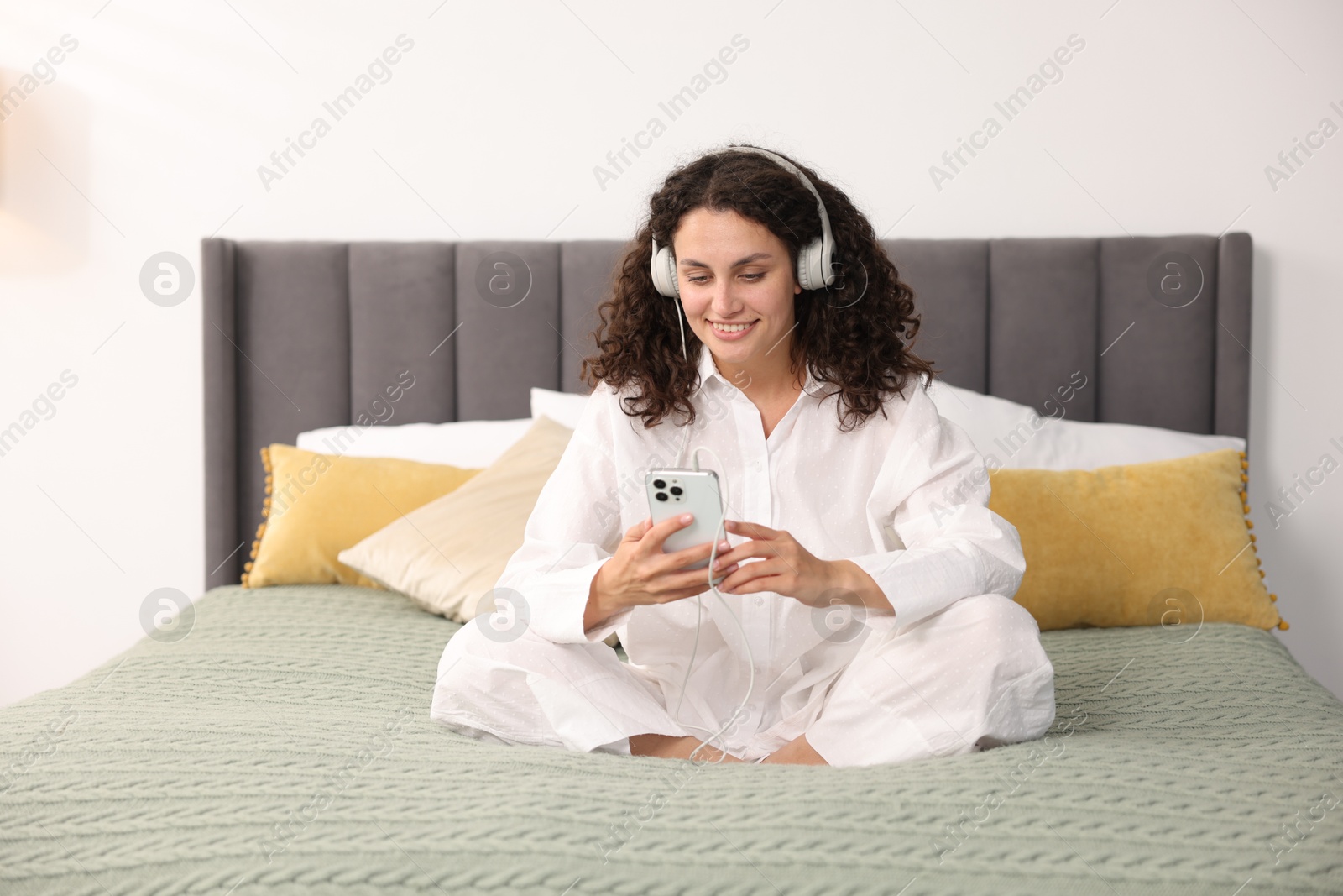 Photo of Young woman with headphones and smartphone listening to music on bed at home