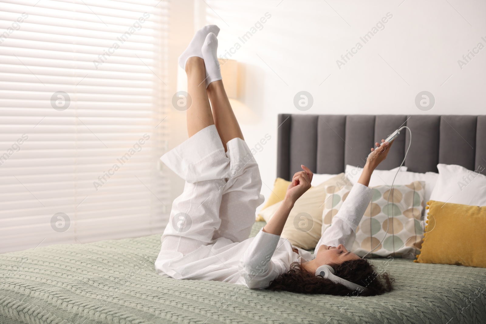 Photo of Young woman with headphones and smartphone listening to music on bed at home