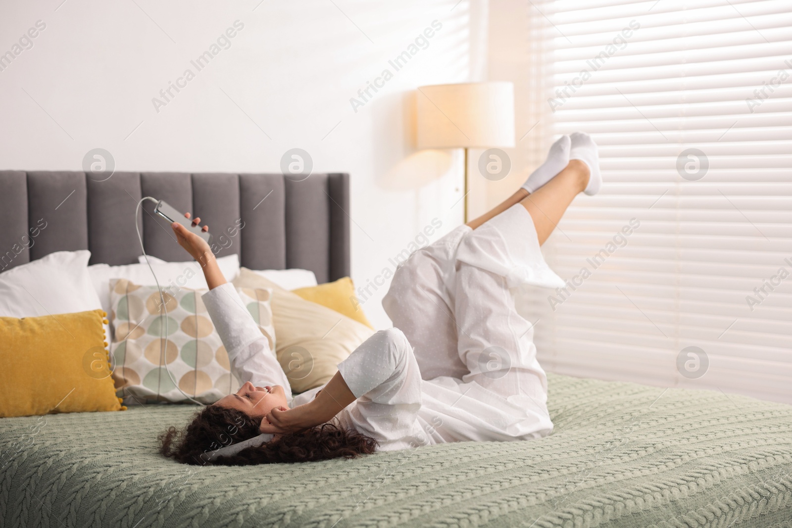 Photo of Young woman with headphones and smartphone listening to music on bed at home