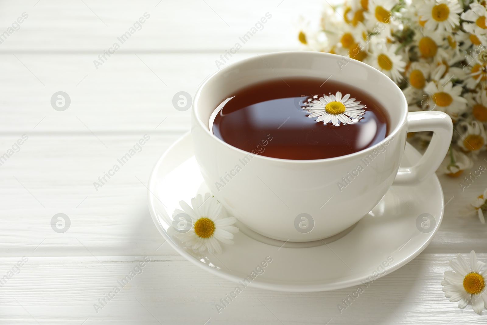 Photo of Aromatic tea in cup and chamomile flowers on white wooden table, closeup