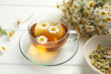 Photo of Aromatic tea in glass cup and chamomile flowers on white wooden table, closeup