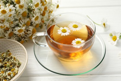 Photo of Aromatic tea in glass cup and chamomile flowers on white wooden table, closeup