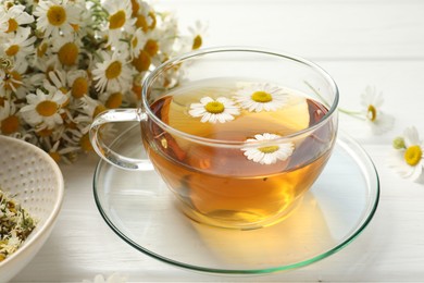 Photo of Aromatic tea in glass cup and chamomile flowers on white wooden table, closeup