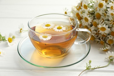 Photo of Aromatic tea in glass cup and chamomile flowers on white wooden table, closeup