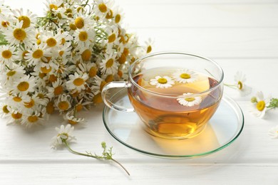 Photo of Aromatic tea in glass cup and chamomile flowers on white wooden table, closeup