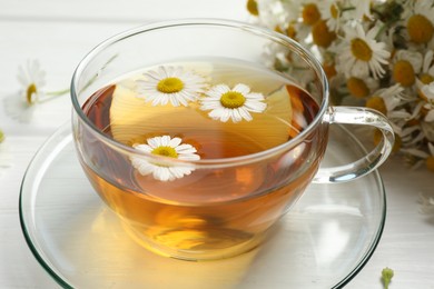 Photo of Aromatic tea in glass cup and chamomile flowers on white table, closeup