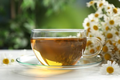Photo of Aromatic tea in glass cup and chamomile flowers on white wooden table, closeup