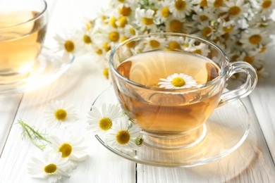 Photo of Aromatic tea in glass cup and chamomile flowers on white wooden table, closeup