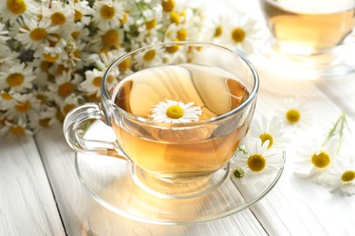 Photo of Aromatic tea in glass cup and chamomile flowers on white wooden table, closeup