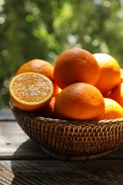 Photo of Fresh whole and cut oranges in wicker basket on wooden table against blurred green background, closeup