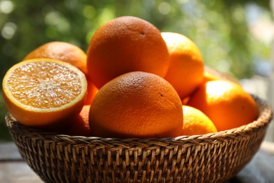 Photo of Fresh whole and cut oranges in wicker basket on wooden table against blurred green background, closeup