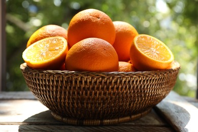Photo of Fresh whole and cut oranges in wicker basket on wooden table against blurred green background, closeup