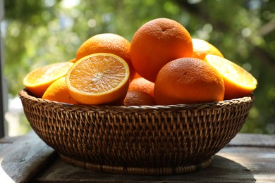 Photo of Fresh whole and cut oranges in wicker basket on wooden table against blurred green background, closeup