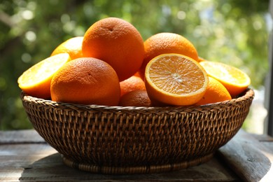 Photo of Fresh whole and cut oranges in wicker basket on wooden table against blurred green background, closeup