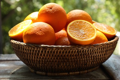 Photo of Fresh whole and cut oranges in wicker basket on wooden table against blurred green background, closeup