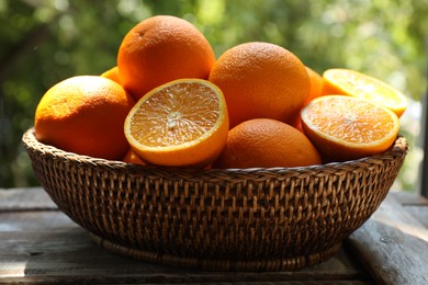 Photo of Fresh whole and cut oranges in wicker basket on wooden table against blurred green background, closeup