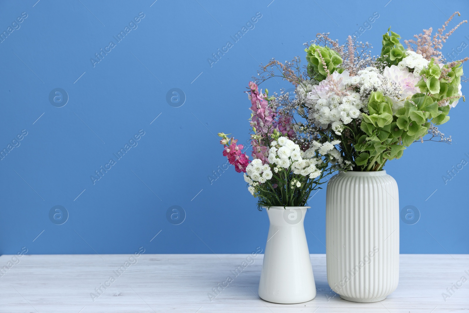 Photo of Beautiful flowers in vases on white wooden table against light blue background, space for text
