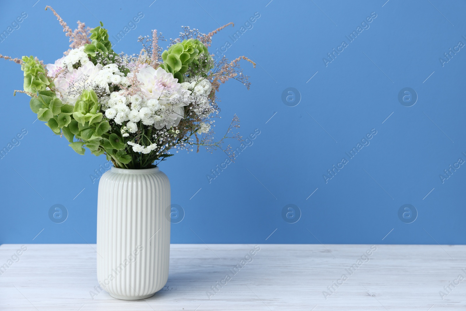 Photo of Beautiful flowers in vase on white wooden table against light blue background, space for text