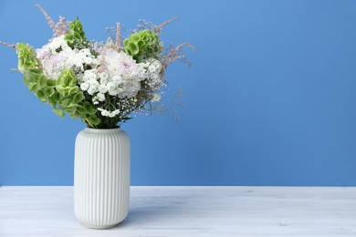 Photo of Beautiful flowers in vase on white wooden table against light blue background, space for text