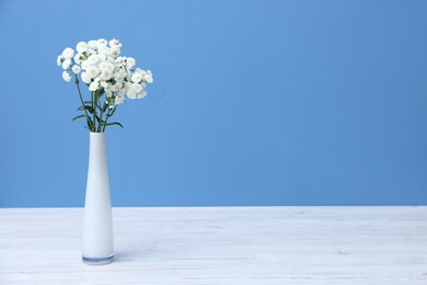 Photo of Beautiful flowers in vase on white wooden table against light blue background, space for text