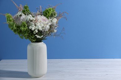 Photo of Beautiful flowers in vase on white wooden table against light blue background, space for text
