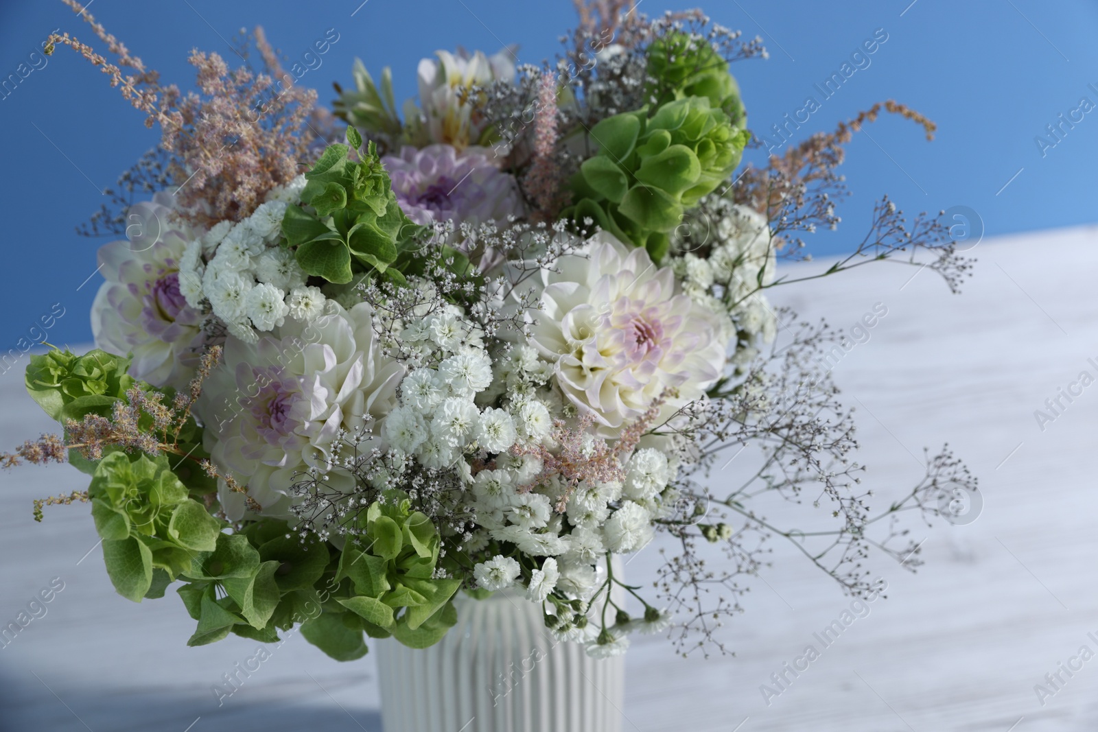 Photo of Beautiful flowers in vase on white wooden table against light blue background, closeup