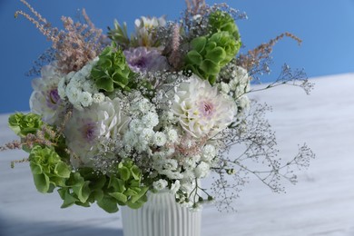 Photo of Beautiful flowers in vase on white wooden table against light blue background, closeup